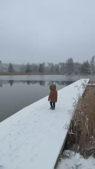 Malé dítě v zimním oblečení kráčí po zasněžené cestě podél klidné vodní hladiny. V pozadí jsou zasněžené stromy a krajina zahalená do zimního počasí. Video zdůrazňuje bezpečnost a klid rodičů díky dětským hodinkám KLOKI na dovolené.
