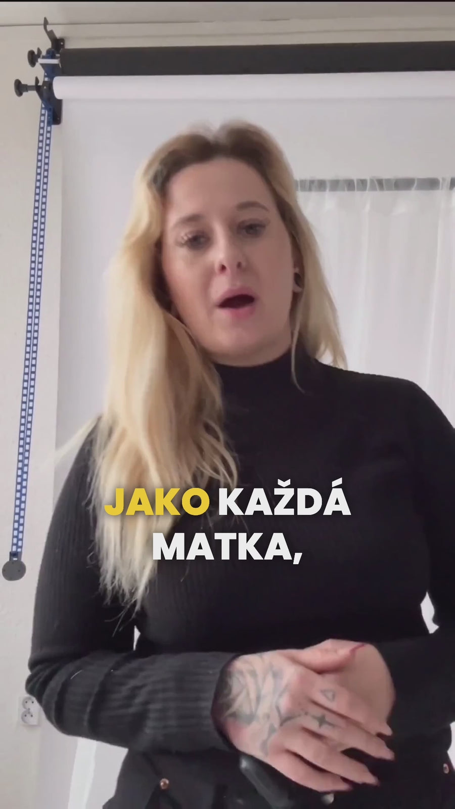 Žena v černém roláku stojí před světlým pozadím a mluví směrem do kamery.  Představuje využití dětských hodinek KLOKI v každodenních situacích.